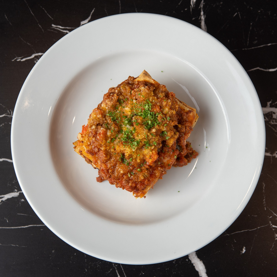 A square of Lexie's Bistro's lasagna, topped with rich meat sauce and fresh parsley, on a white plate.