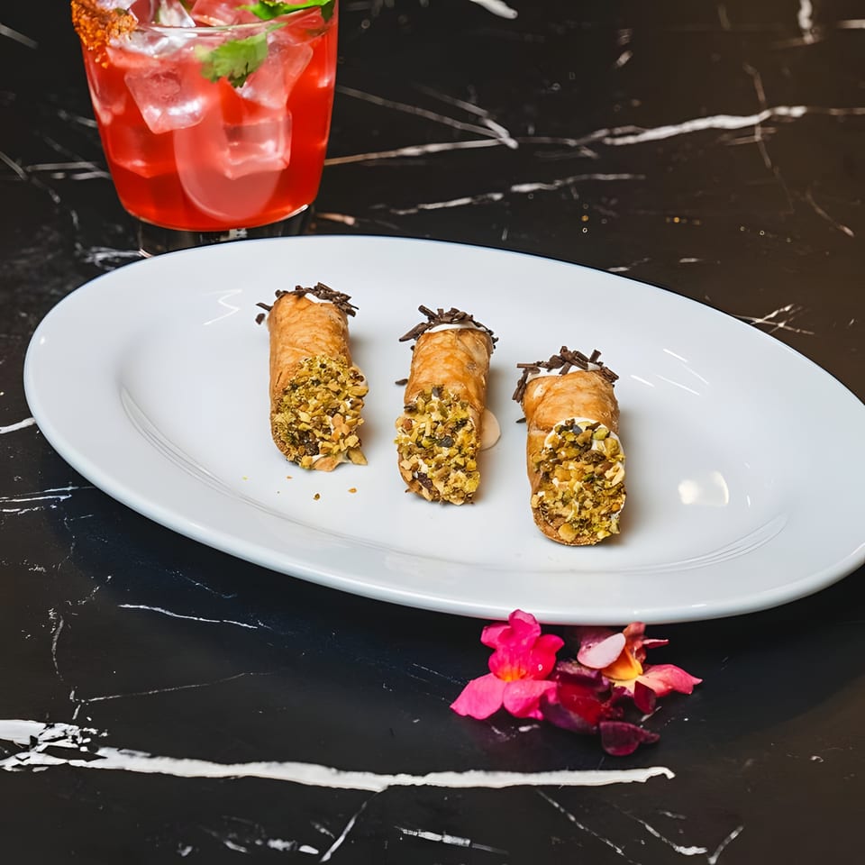 Three Lexie's Bistro cannolis with pistachios and chocolate shavings, next to a red cocktail.